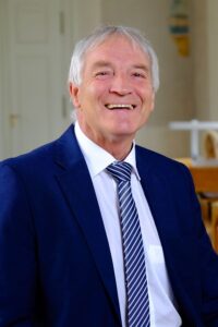 An elderly man in a blue suit and tie smiling while sitting in a bright room