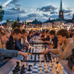 A large group of people play chess at long tables on the waterfront in the evening, with string lights glowing around them and city lights and a festive open-air chess festival atmosphere visible in the background.
