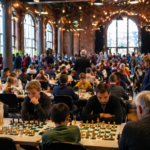 A large chess festival takes place in a spacious hall with tall windows and strings of lights, where participants of different ages are playing at many tables, while a lively atmosphere of a major chess event fills the space.