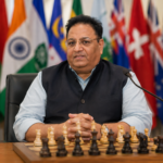 Ο Bharat Singh Chauhan κάθεται σε σκακιέρα με φόντο σημαίες χωρών της Κοινοπολιτείας κατά τη διάρκεια διεθνούς σκακιστικής διοργάνωσης.