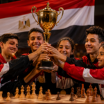 Grupo de adolescentes con uniformes deportivos rojos y negros sosteniendo un trofeo dorado y sonriendo durante una ceremonia de premiación en un torneo de ajedrez; la bandera de Egipto aparece al fondo y un tablero de ajedrez está en primer plano.