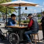 Outdoor scene in Trelleborg, Sweden showing a mobile chess station mounted on a bicycle cart. An older man plays chess against a teenager under a large umbrella while several young spectators watch in a sunny waterfront park.